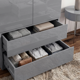 [Grey] Close view of high gloss wardrobe drawers open with folded clothes, books, towels, and dark interior.