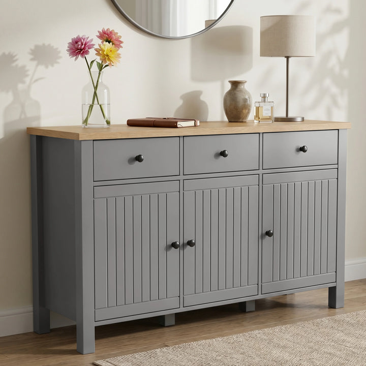 [Grey] Linra angled front view with three drawers, beadboard doors, oak-effect top, mirror, lamp, and flowers