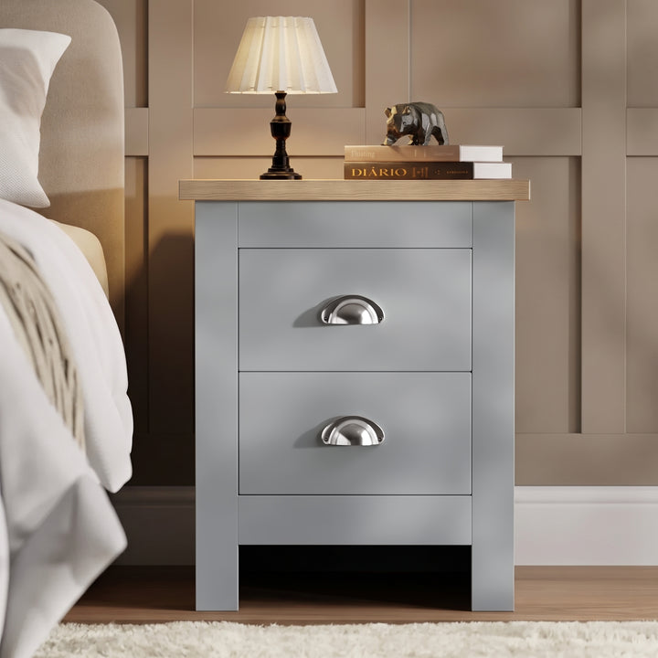 [Grey] Bedside table front view beside a bed, with lamp, stacked books, panelled wall, and shaggy rug