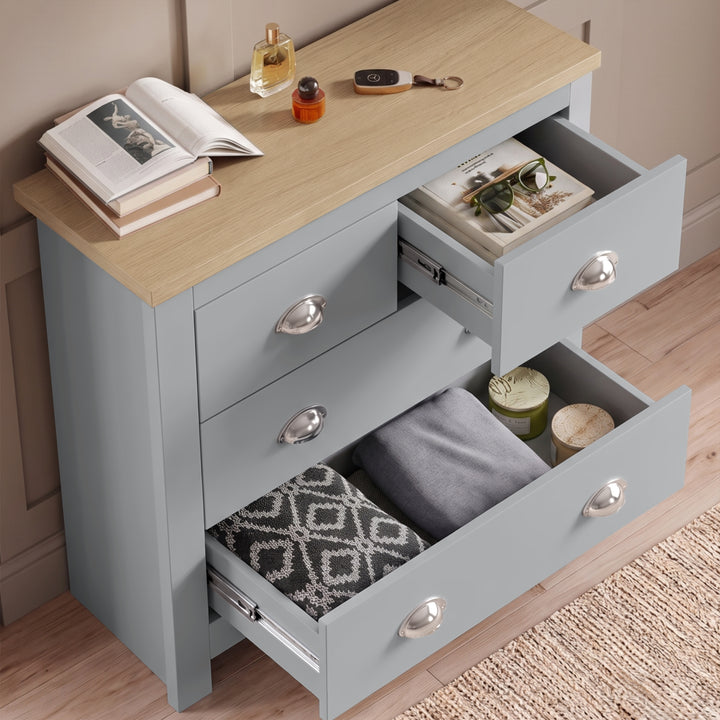 [Grey] Chest of drawers angled view with two drawers open, folded textiles, tray, book, perfume, and oak top