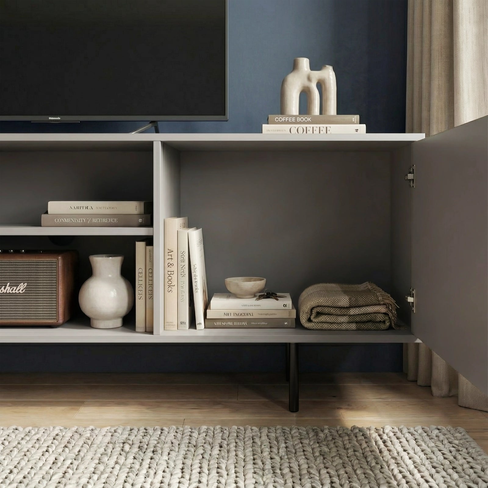 [Grey] Open-door view of grey TV unit, internal compartments holding books and home accessories, dark navy wall behind.