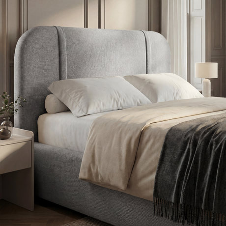 Grey fabric headboard with curved top edge and padded surface, styled with layered bedding in neutral tones