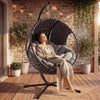 [Dark Grey] A round dark grey hanging egg chair in a cozy garden setting, offering comfort and relaxation to the girl seating inside.