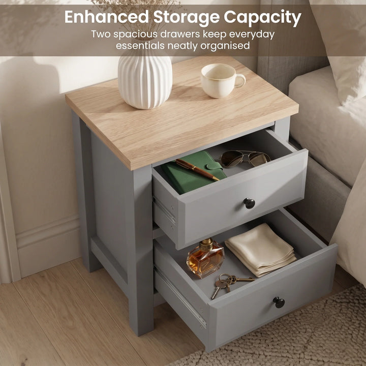 [Grey] two-drawer nightstand with a light wood top, shown beside a bed in a bedroom. Two drawers are open, displaying storage space with small items inside.