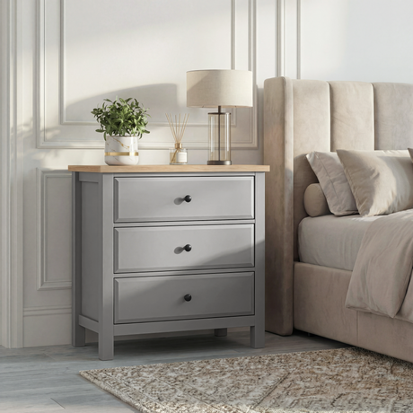 [Grey] Haven 3-drawer chest in a bedroom setting with lamp on oak top and upholstered headboard panel behind
