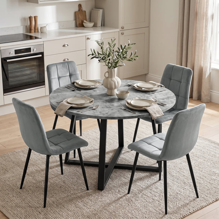 [Light Grey]  A round gray stone-look dining table with a black metal base, surrounded by four gray upholstered chairs, set in a bright modern kitchen with light wood floors and beige curtains.