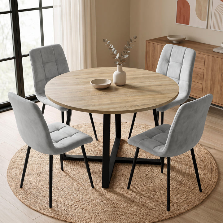 [Light Grey | Sonoma Oak] A round light oak dining table with a black metal pedestal base, surrounded by four light gray tufted velvet chairs with black legs, set on a round woven jute rug in a modern dining room.