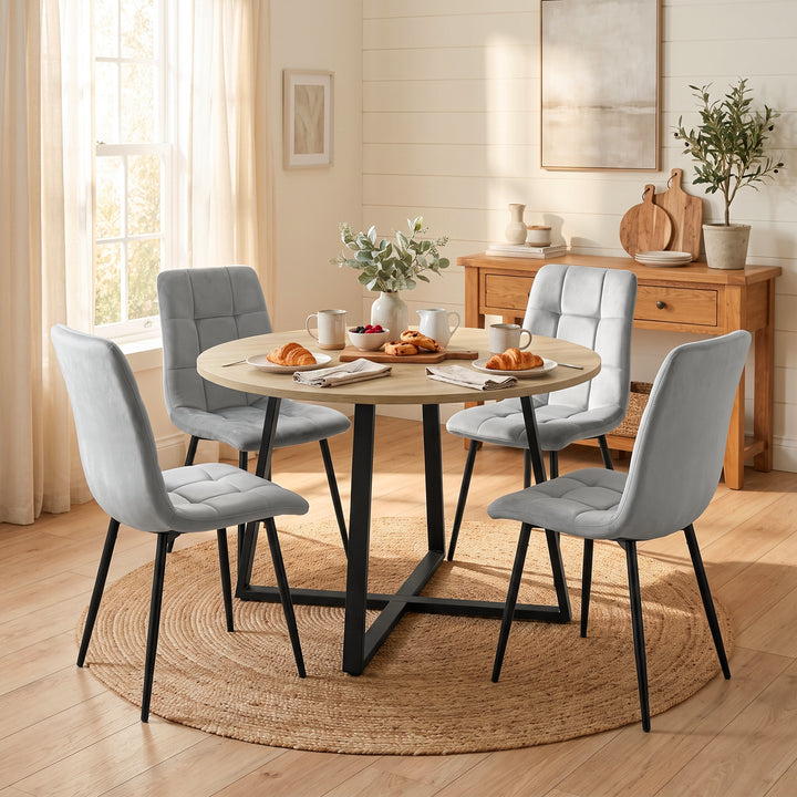 [Light Grey | Sonoma Oak] A 5-piece dining set featuring a round light wood table with a black metal X-frame base and four tufted light gray velvet chairs with black legs, styled in a bright, modern dining room.