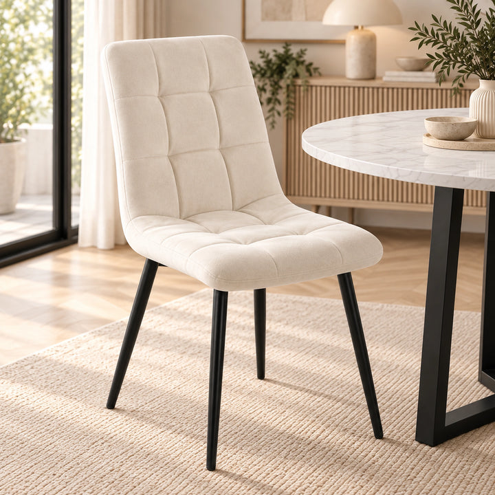 [Cream] A modern dining chair with a cream tufted upholstered seat and back, supported by slim black metal legs, shown in a bright, contemporary dining room next to a marble-top table.