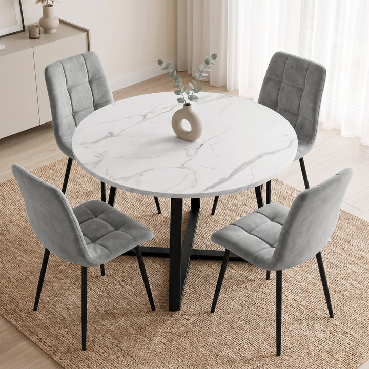 [Light Grey | White Marble] A round dining table with a white marble-patterned top and black metal legs, surrounded by four gray upholstered chairs with black legs, set on a woven rug in a bright dining room.