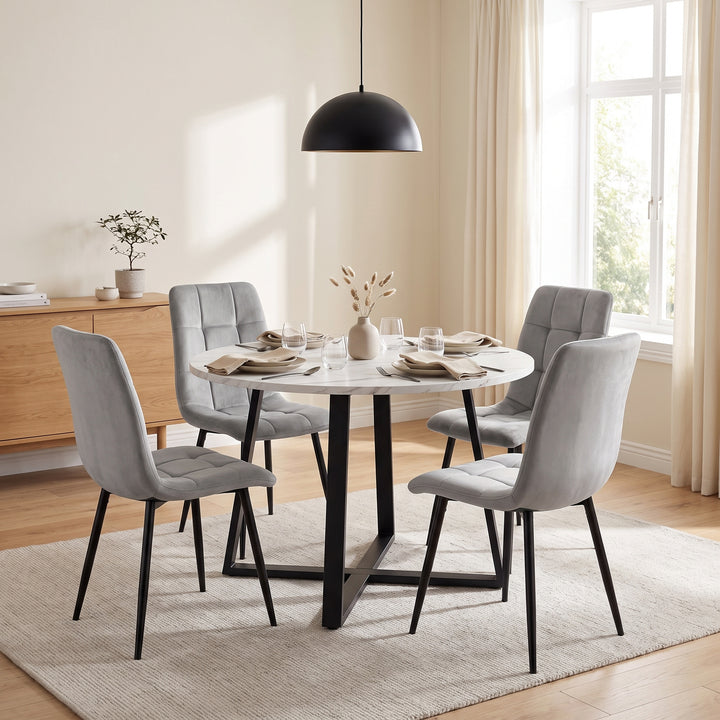 [Light Grey | White Marble] A round white dining table with a black metal base, surrounded by four light gray upholstered chairs with black legs, set on a beige rug in a bright dining room.