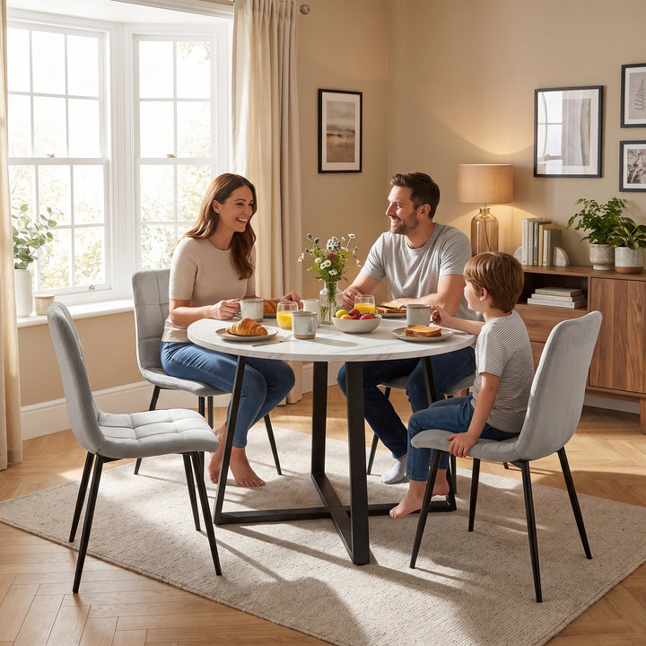 [Light Grey | White Marble] A family of three shares a meal at a round white dining table with a black metal base, surrounded by light gray upholstered chairs in a warm, modern dining room.