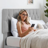 Woman relaxing on a light grey double bed frame with a tufted headboard in a cozy bedroom.