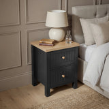 [Black] Bedroom view of the Linra bedside table by a linen bed with a jute rug and panelled wall behind