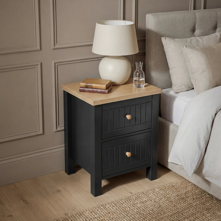 [Black] Bedroom view of the Linra bedside table by a linen bed with a jute rug and panelled wall behind