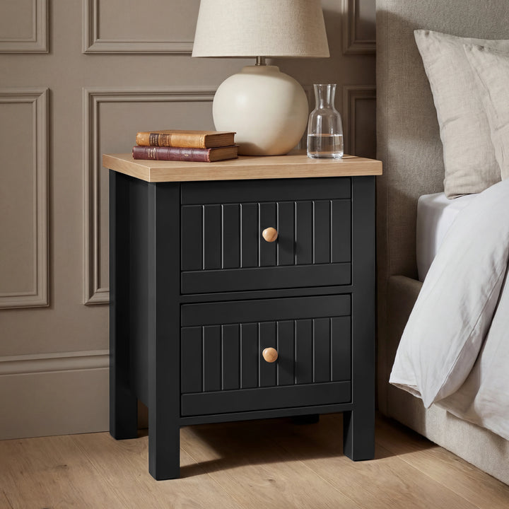 [Black] Black Linra bedside table with two grooved drawers and round wooden knobs styled next to a cream linen bed