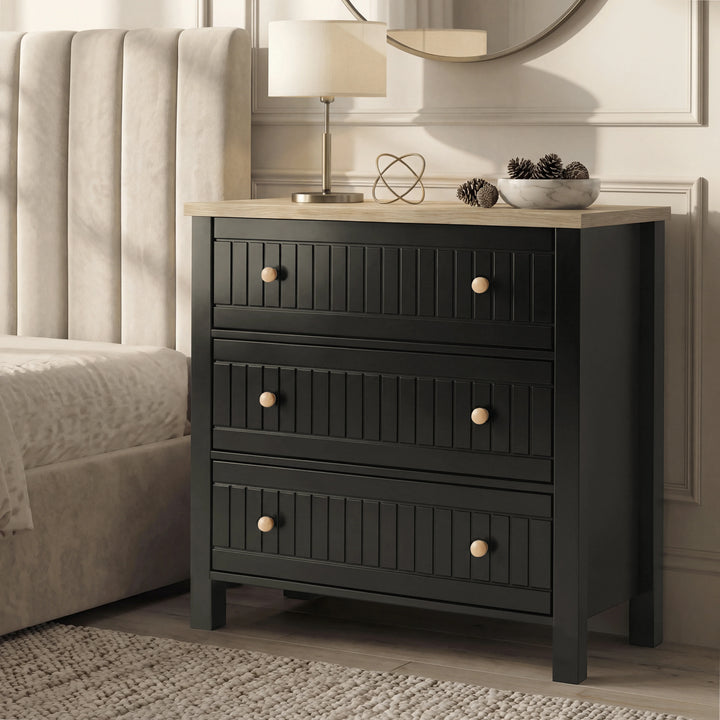 [Black] Linra chest of drawers in black at side of panelled bedroom wall, lamp and pinecones on oak top