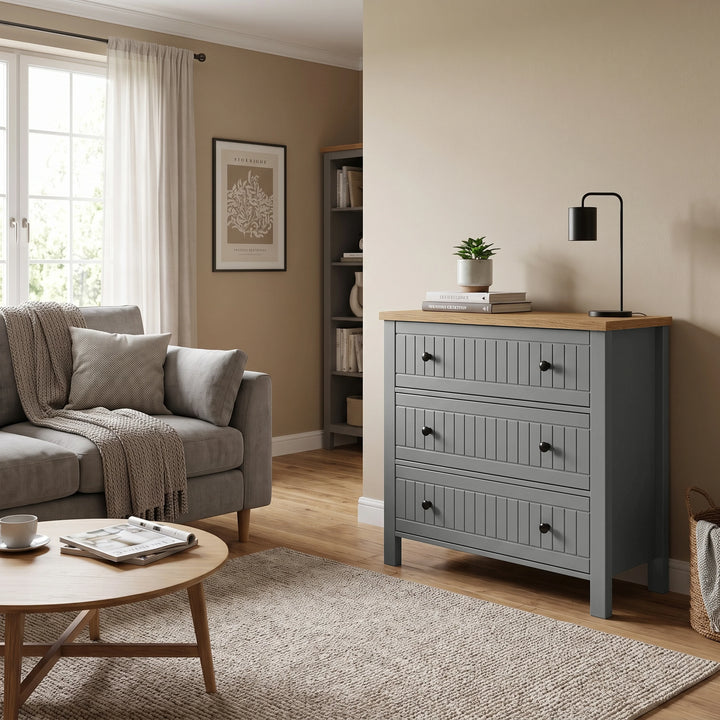 [Grey] Linra 3-drawer chest styled as living room storage beside a sofa, with black lamp, plant and books on the oak veneer top