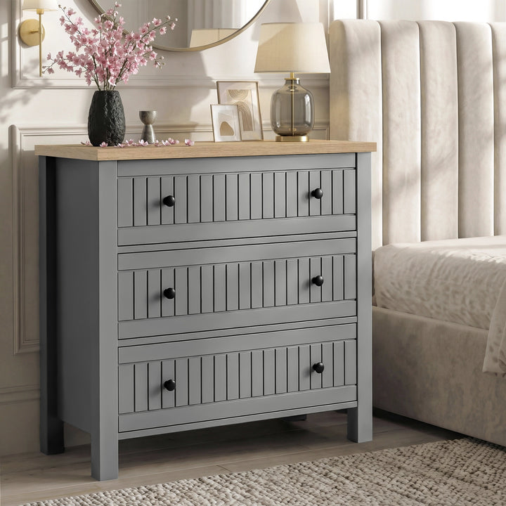 [Grey] Linra 3-drawer chest used as a bedside table in a bedroom with lamp and accessories on oak veneer top