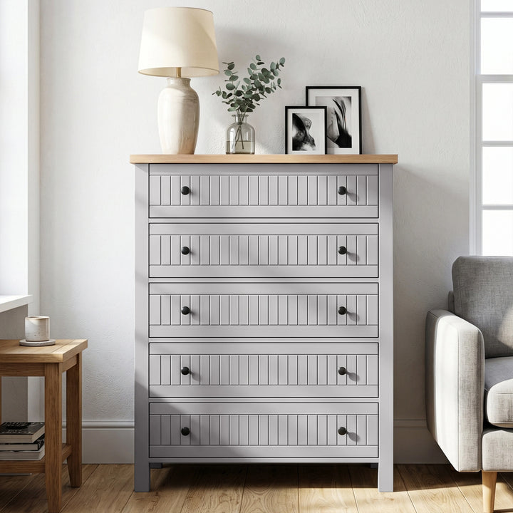 [Grey] Linra 5-drawer chest front view in a neutral styled room with lamp and decorative objects on oak veneer top