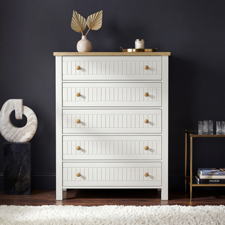 [White] Linra 5-drawer chest front view in a styled room with dark wall and decorative objects on oak veneer top