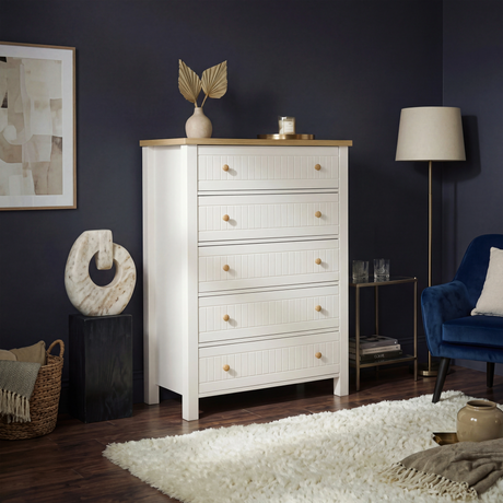 [White] Linra 5-drawer chest front view in a styled bedroom with floor lamp and decorative objects on oak top