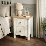 [White] Linra 2-drawer bedside table in a bedroom lifestyle setting with soft furnishings and lamp in background