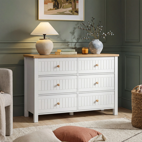 [White] Linra 6-drawer chest angled view in a bedroom with dark wall, lamp on oak top and artwork in background