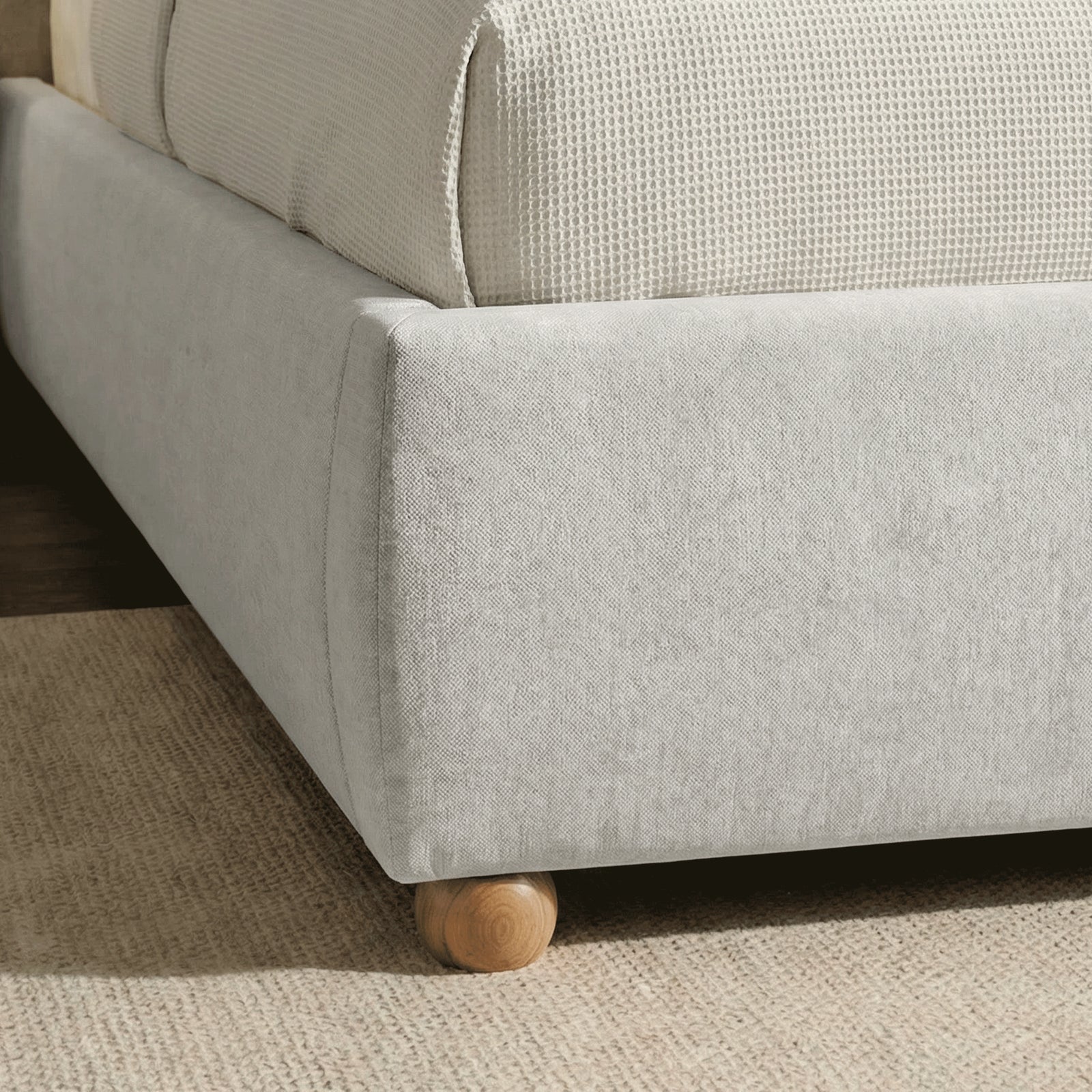 [Grey | King] Bed frame foot detail showing single round wooden leg and padded base with textured throw visible above