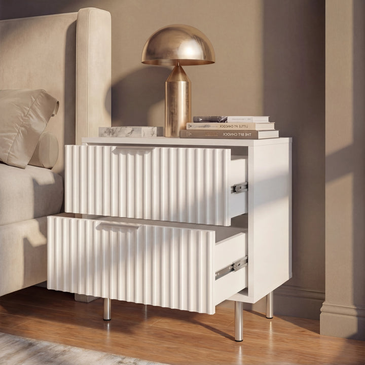 [White] bedside table with silver legs and partially opened drawers, placed against a wall in a naturally lit bedroom