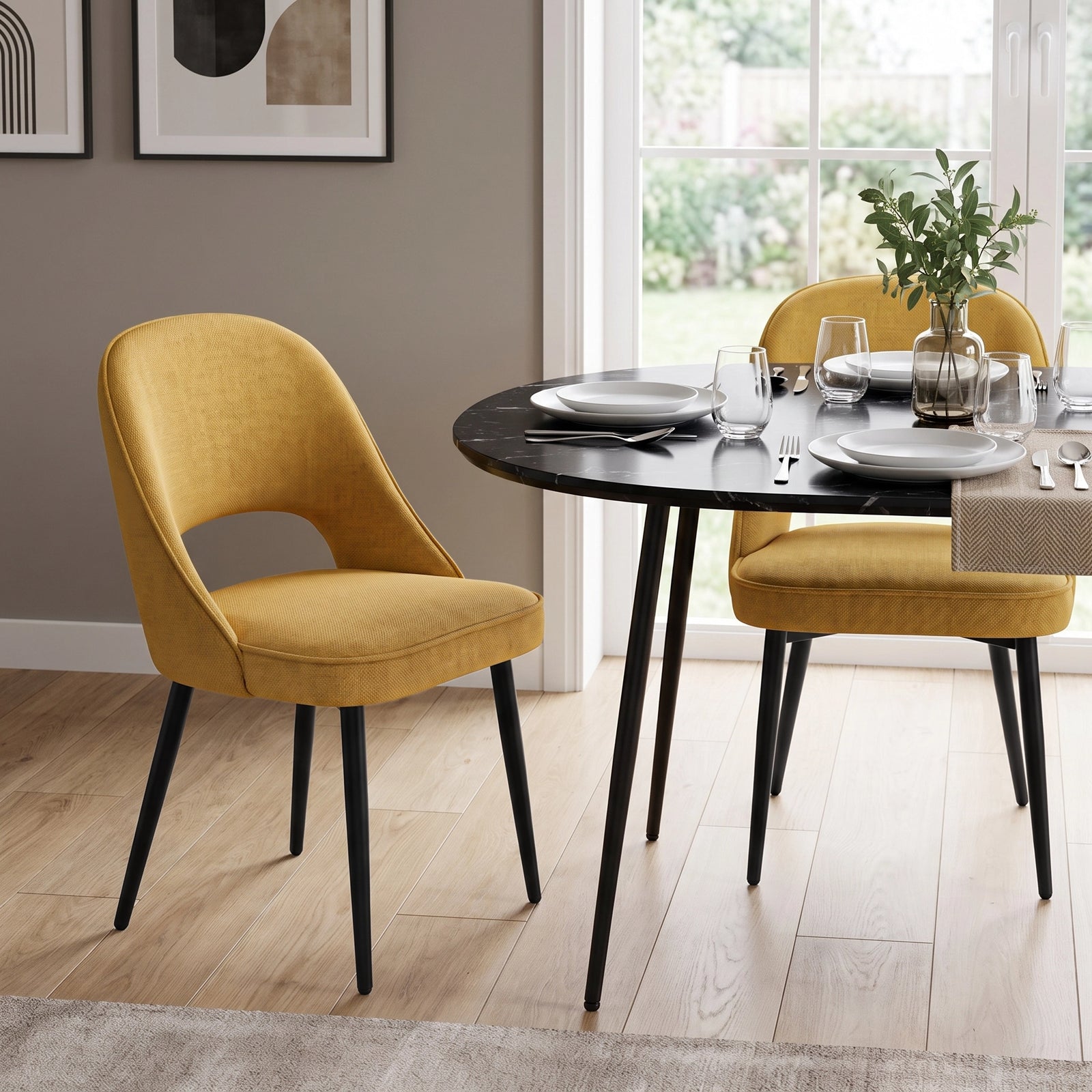 [Mustard | Set of 2] Pair of curved-back comfy dining chairs around a round black table with plates, glasses & a vase.