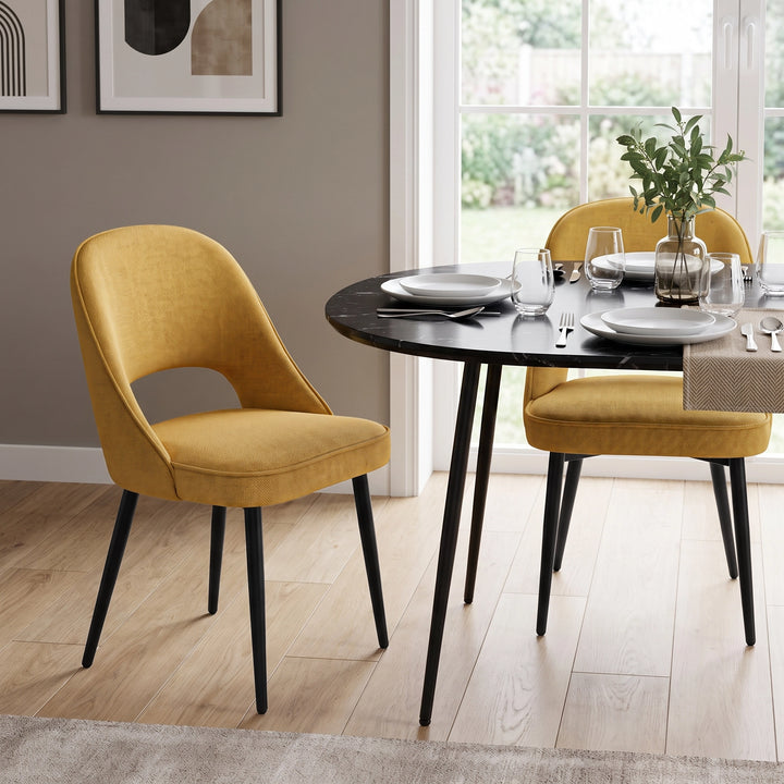 [Mustard | Set of 2] Pair of curved-back comfy dining chairs around a round black table with plates, glasses & a vase.