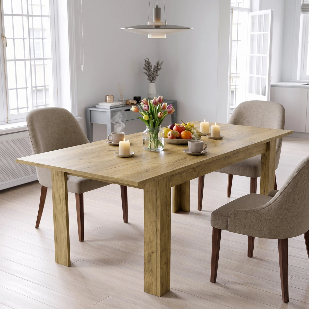 [Oak] Closed oak-effect dining table styled with fruit, flowers and candles; beige chairs stand by its thick legs.