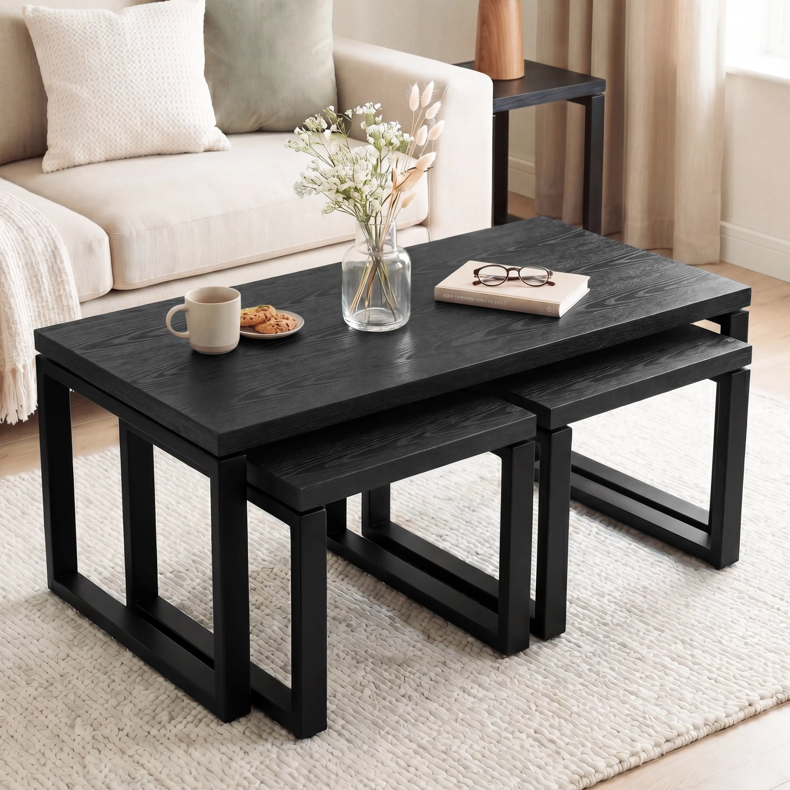[Black] Nestline coffee table set beside a cream sofa, with vase flowers, book, cup, and black nesting frames