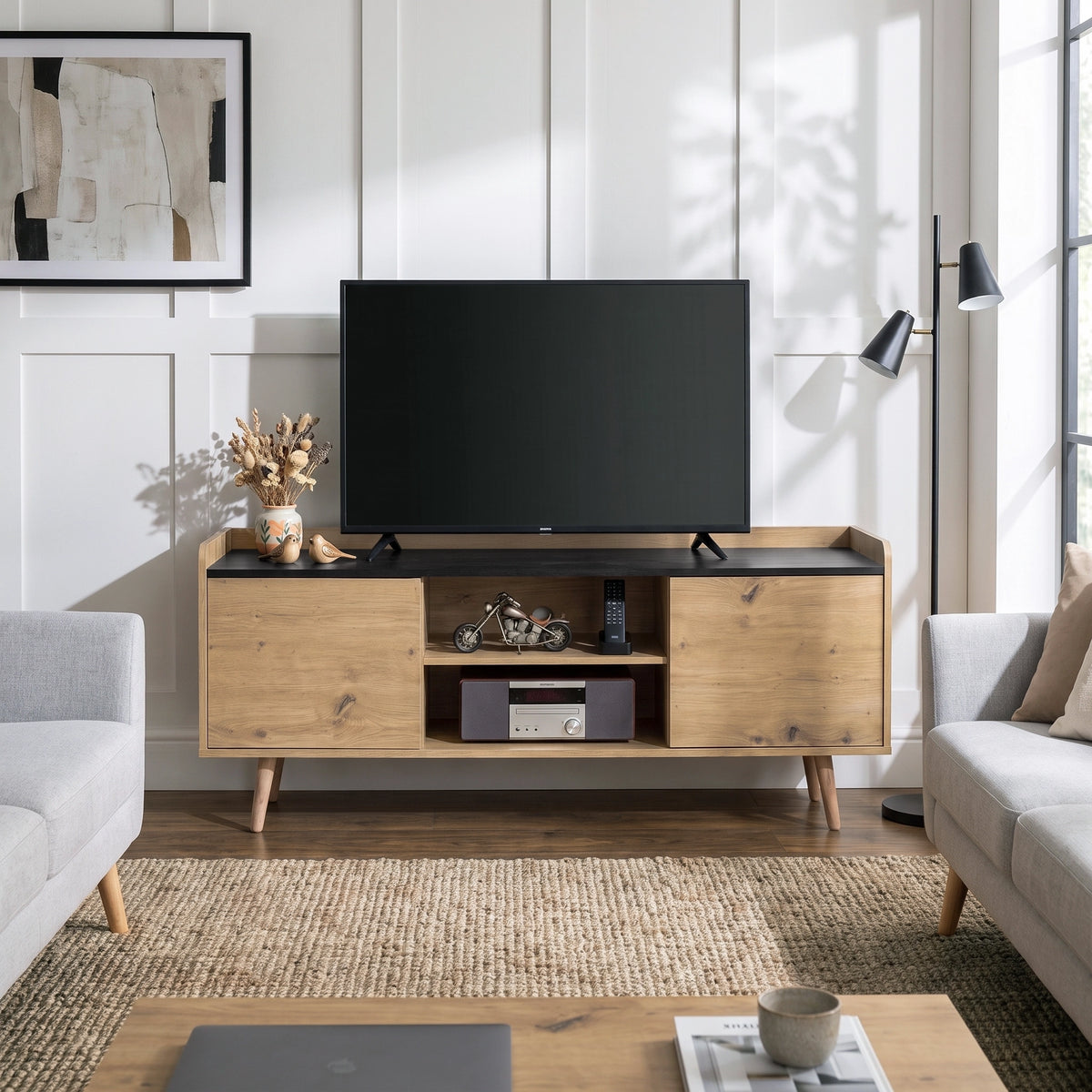 [Oak] A mid-century modern TV stand console in light natural wood with a black top and tapered legs, styled in a bright living room with a flat-screen TV, sofa, and rug.