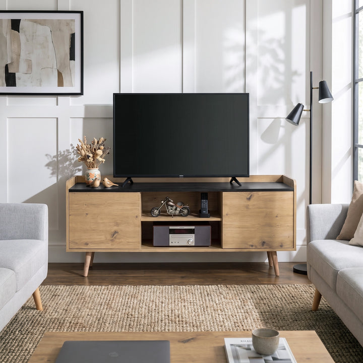 [Oak] A mid-century modern TV stand console in light natural wood with a black top and tapered legs, styled in a bright living room with a flat-screen TV, sofa, and rug.