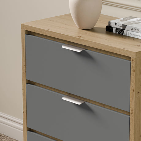 Oak and grey finished chest of drawers, with metallic handles, placed against a wall with a vase, and books on the top