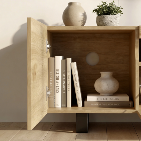 [Oak] Kystra TV stand left cabinet door open close-up, showing internal shelf with books and a ceramic vase, chrome hinge hardware, and a round cable routing hole in the back panel
