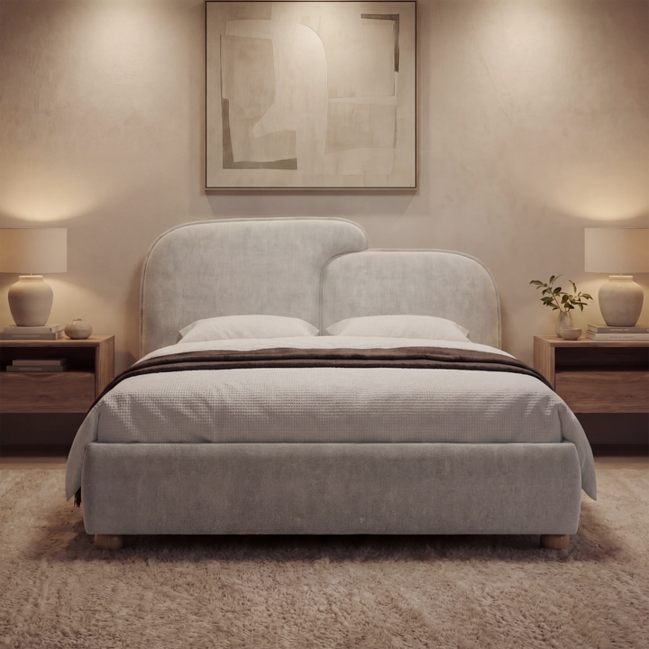 [Double | Grey] Grey fabric double ottoman bed frame centered under framed art with wooden nightstands and lamp light