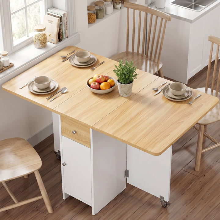 [Pine] A light wood and white foldable dining table with attached storage cabinet and wheels, shown in a bright kitchen setting with chairs, tableware, fruit, and a small plant.