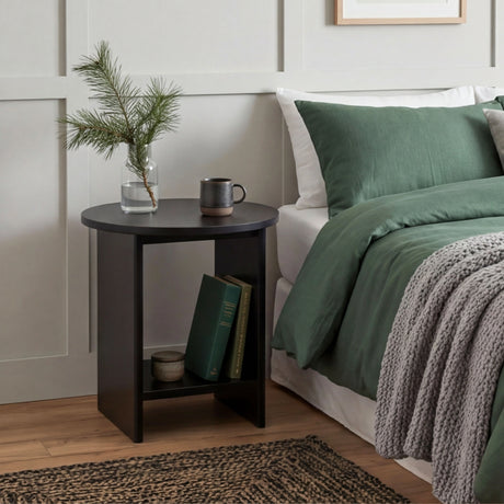 [Black] Black round side table used as bedside next to green duvet bed, glass vase pine sprig and mug on top