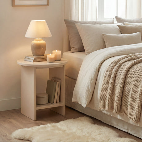 [Taupe] Round taupe end table beside bed dressed in chunky cream knit throw, lamp and books on top