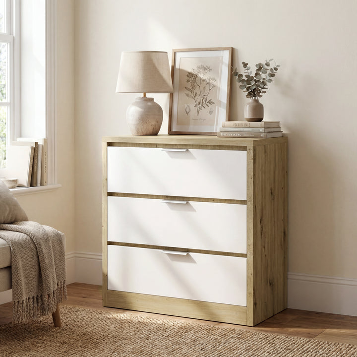[Oak+White] Side-angled Skova chest of drawers next to cream armchair with throw, lamp glowing on oak top