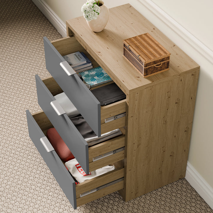[Oak+Grey] Angled view of Skova chest, three grey drawers stepped open revealing towels, folded items and a red garment