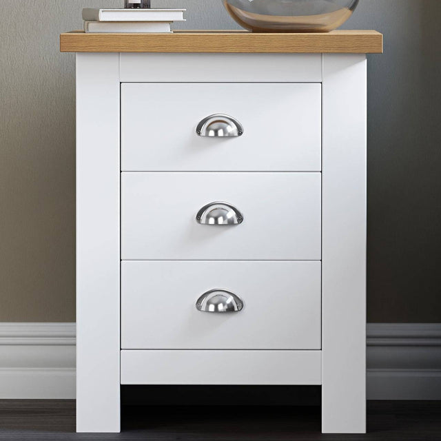 Small white bedside table with handles, featuring a cozy top surface with a lamp and books.