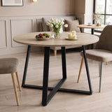 [Sonoma Oak] Round light oak-effect dining table in a bright room with flowers, fruit and candles on the tabletop.
