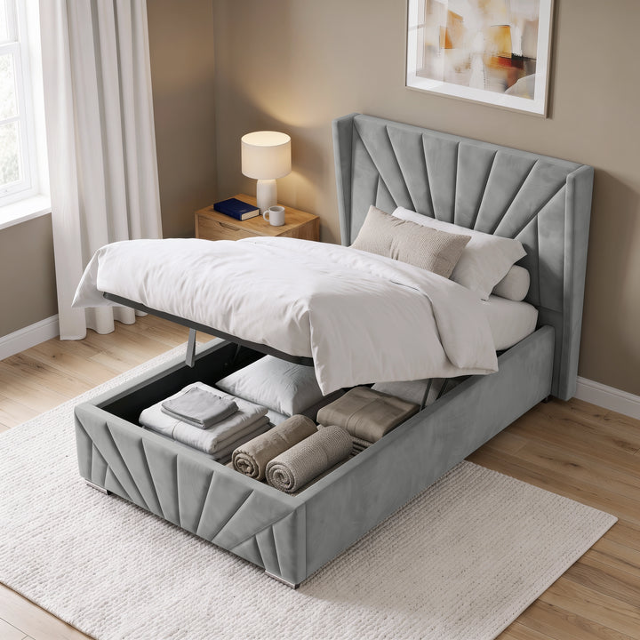 [Single | Grey] upholstered bed with a tall tufted headboard and raised storage base, shown in a styled bedroom with white bedding, folded towels, a nightstand, lamp, curtains, and wood flooring.