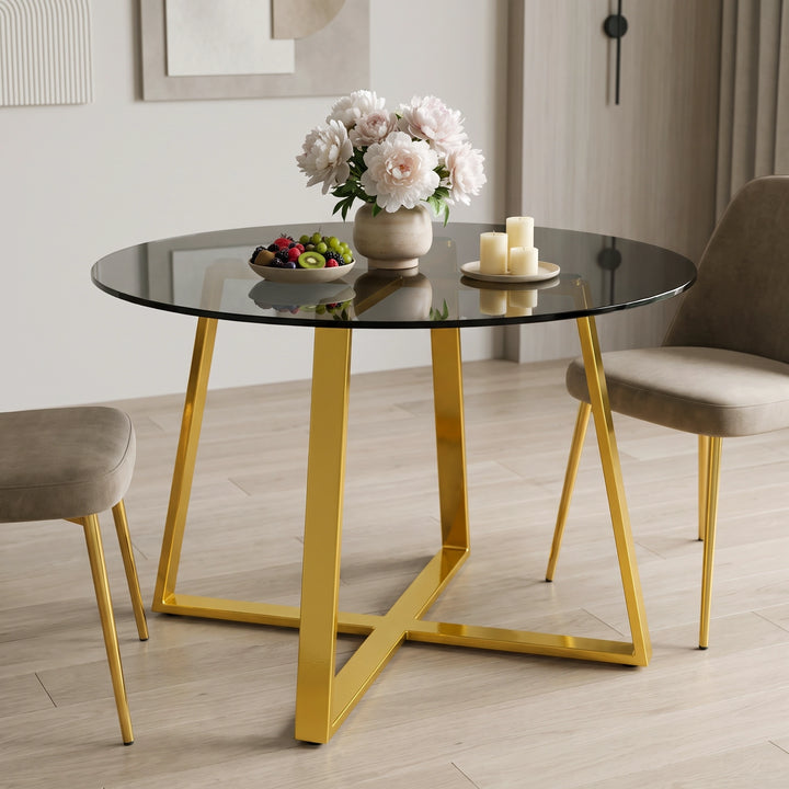 [Tinted] round dining table with a tinted glass top and a polished gold metal base, styled in a modern dining room with two beige upholstered chairs, a vase of flowers, and candles.