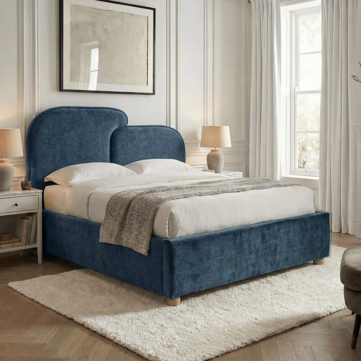 Double bed upholstered in blue fabric placed near a window with light bedding and neutral bedroom decor