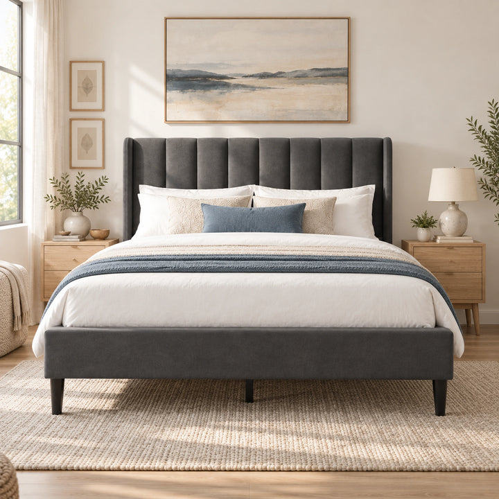 A modern upholstered bed frame in dark gray velvet, styled in a bright, neutral-toned bedroom with white bedding, a blue accent pillow, wooden nightstands, and a large landscape painting above the headboard.